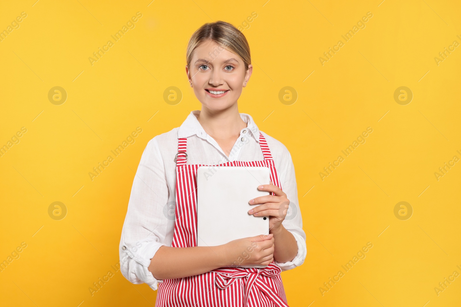 Photo of Beautiful young woman in clean striped apron with tablet on orange background