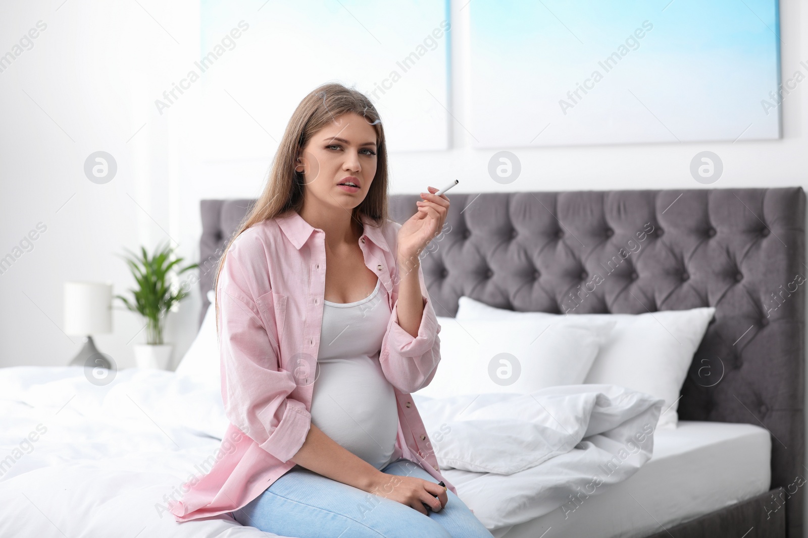 Photo of Young pregnant woman smoking cigarette in bedroom. Harm to unborn baby