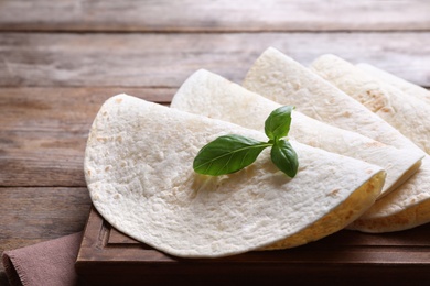 Photo of Board of tasty tortillas with basil leaves on wooden table, closeup