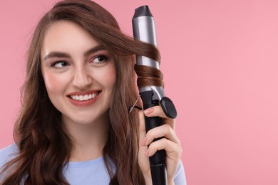 Photo of Beautiful young woman using curling hair iron on pink background, space for text