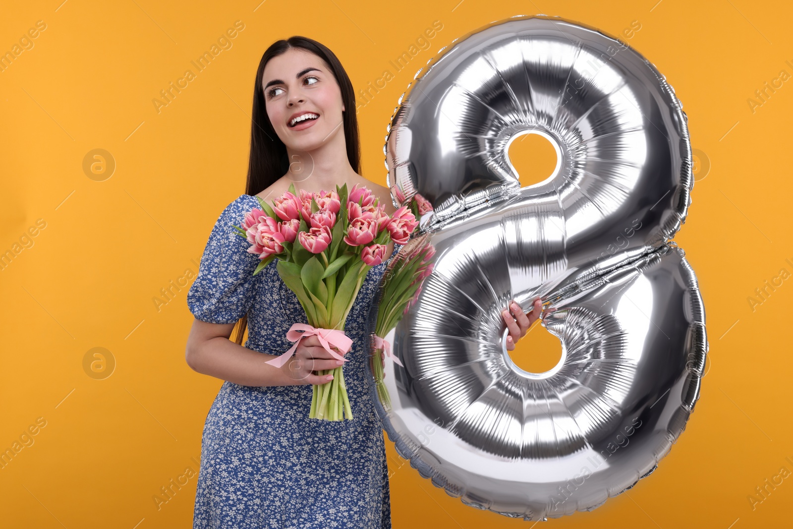 Photo of Happy Women's Day. Charming lady holding bouquet of beautiful flowers and balloon in shape of number 8 on orange background