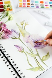 Photo of Woman painting eustomas in sketchbook at white table, closeup