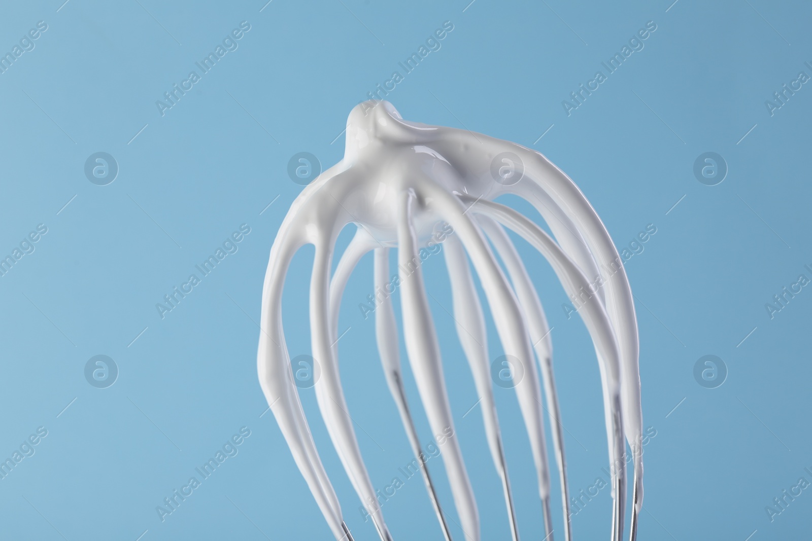 Photo of Whisk with whipped cream on light blue background, closeup