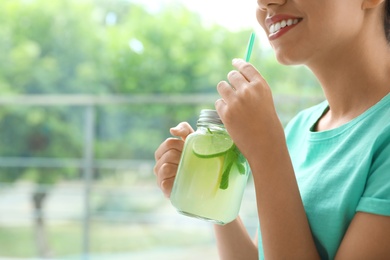 Photo of Happy African-American woman with mason jar of natural lemonade near window indoors, closeup. Detox drink