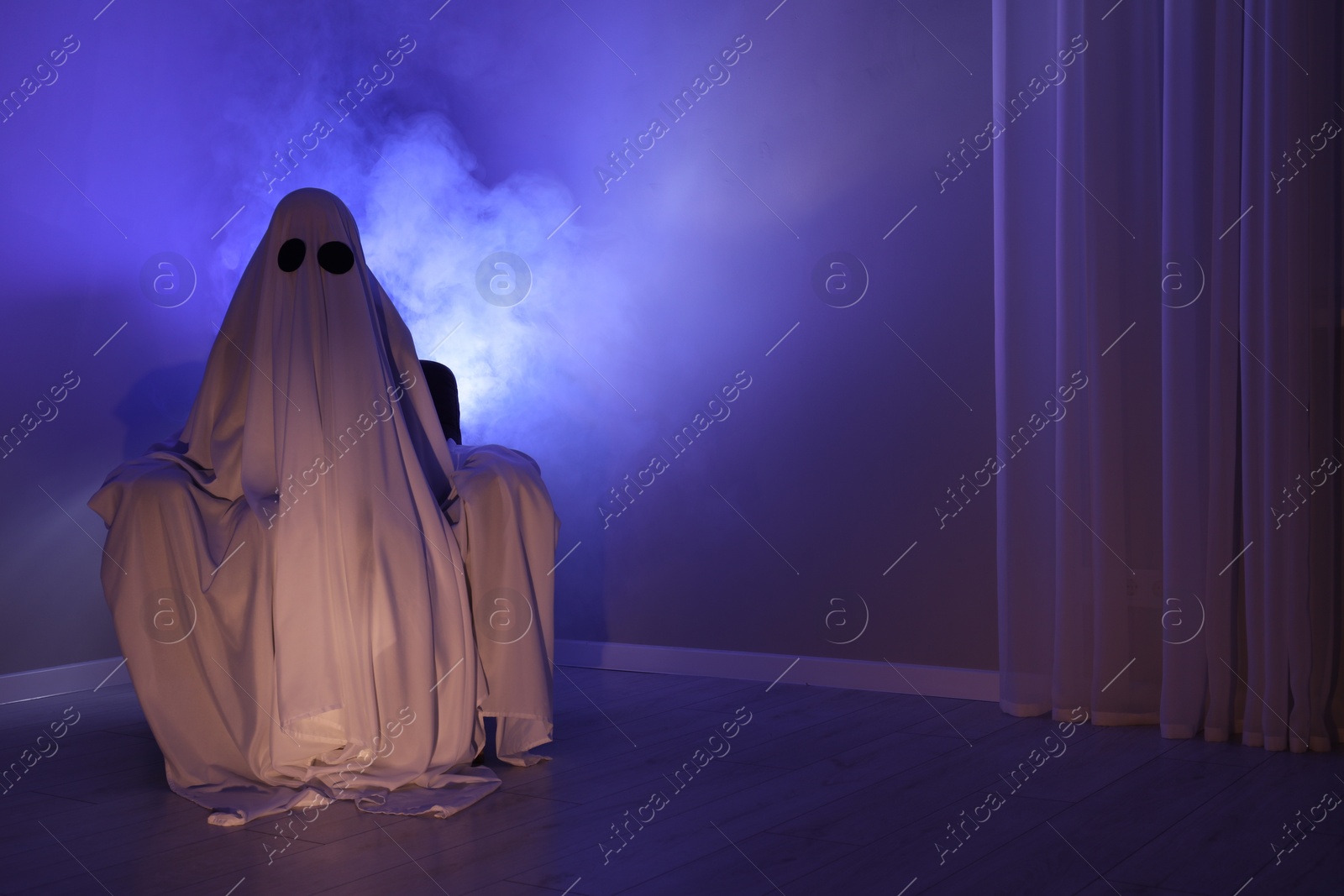 Photo of Creepy ghost. Woman covered with sheet sitting in armchair in blue light, space for text