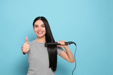 Beautiful happy woman showing thumbs up while using hair iron on light blue background. Space for text