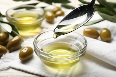 Spoon with cooking oil over bowl and olives on white wooden table, closeup