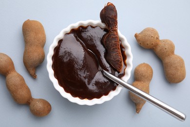 Photo of Bowl of tamarind sauce, spoon and fresh pods on light grey table, flat lay