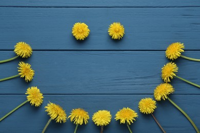 Funny face made of beautiful yellow dandelions on blue wooden table, flat lay