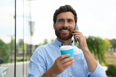 Handsome man with cup of coffee talking on phone outdoors