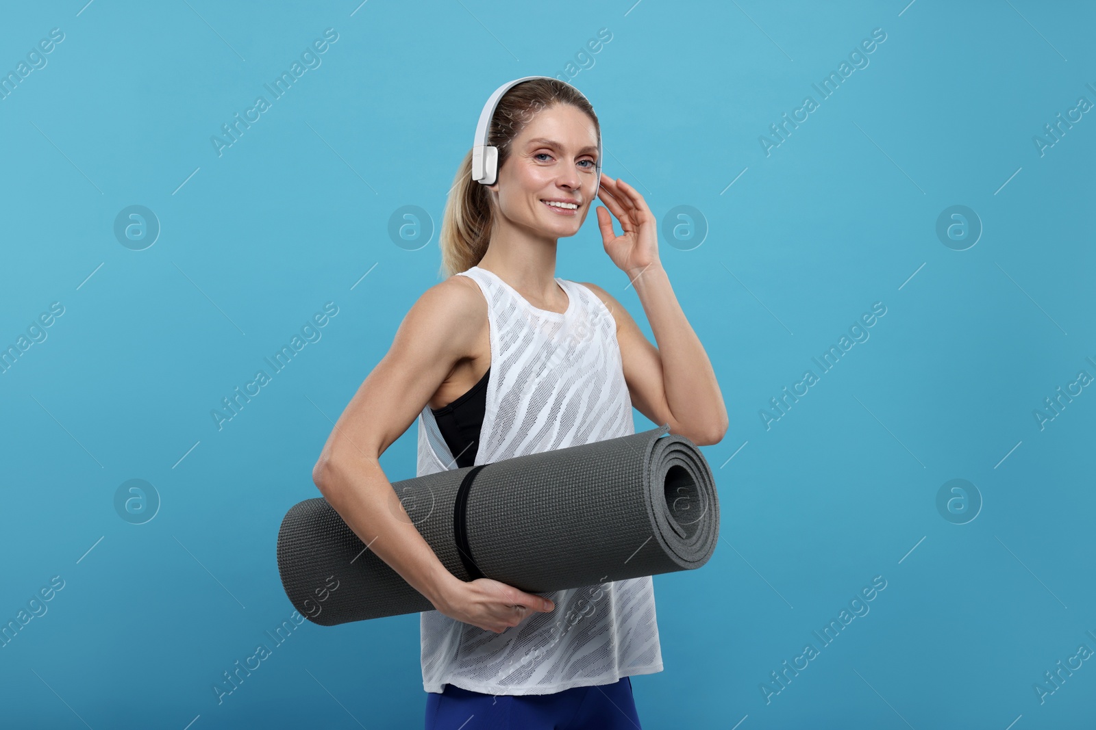 Photo of Sportswoman with headphones and fitness mat on light blue background, space for text