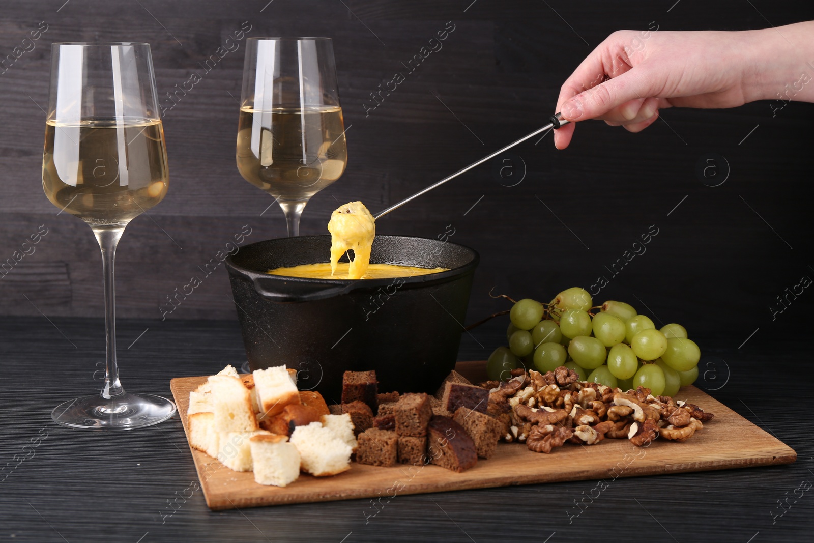 Photo of Woman dipping piece of bread into fondue pot with melted cheese at black wooden table, closeup