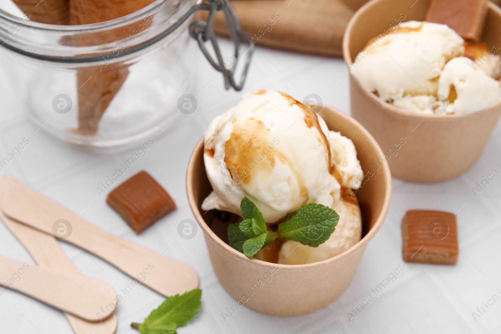 Photo of Scoops of ice cream with caramel sauce, mint leaves and candies on white tiled table, closeup