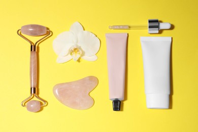 Photo of Gua sha stone, face roller, cosmetic products and orchid flower on yellow background, flat lay
