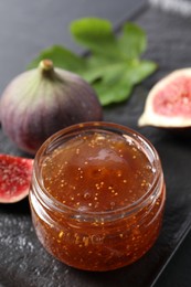Photo of Jar of tasty fig jam and fresh fruits on black board, closeup