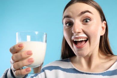 Emotional woman with milk mustache holding glass of tasty dairy drink on light blue background