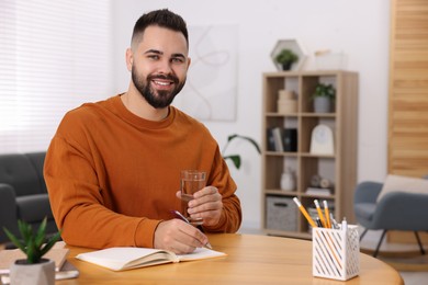 Photo of Young man with glass of water writing in notebook at wooden table indoors