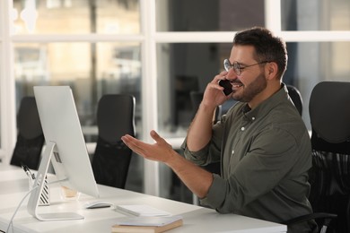 Happy man using modern computer while talking on smartphone at white desk in office