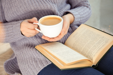 Woman with cup of coffee reading book at home, closeup