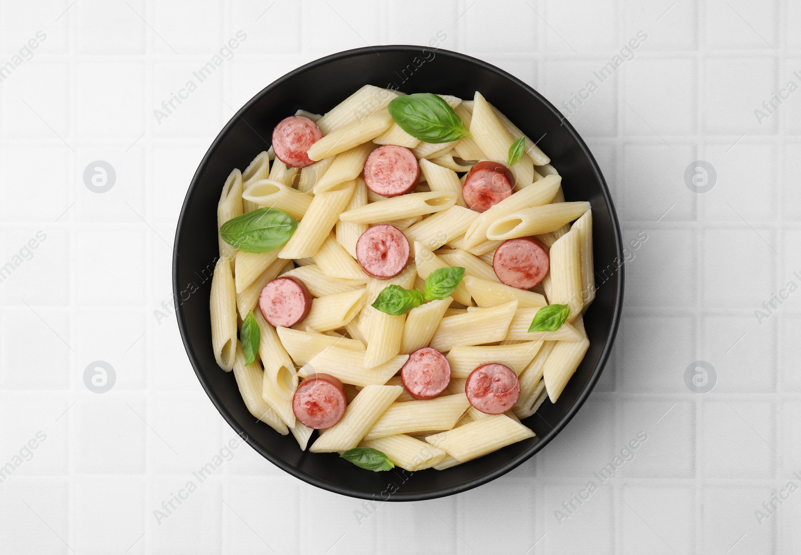 Photo of Tasty pasta with smoked sausage and basil in bowl on white tiled table, top view