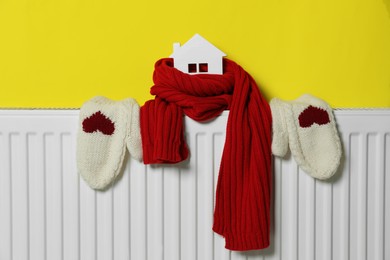 Photo of Wooden house model, knitted scarf and mittens on heating radiator near yellow wall. Energy efficiency concept