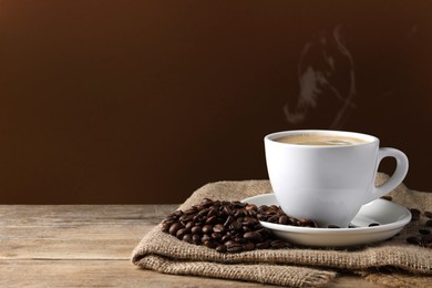 Image of Cup of hot aromatic coffee and roasted beans on wooden table against brown background. Space for text