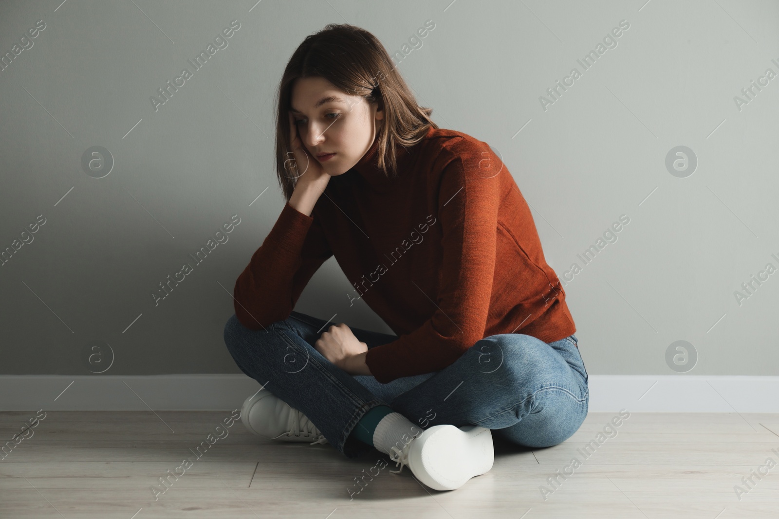 Photo of Sad young woman sitting on floor near grey wall indoors