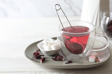 Delicious hibiscus tea in cup, sugar cubes and dry roselle petals on white wooden table, closeup. Space for text
