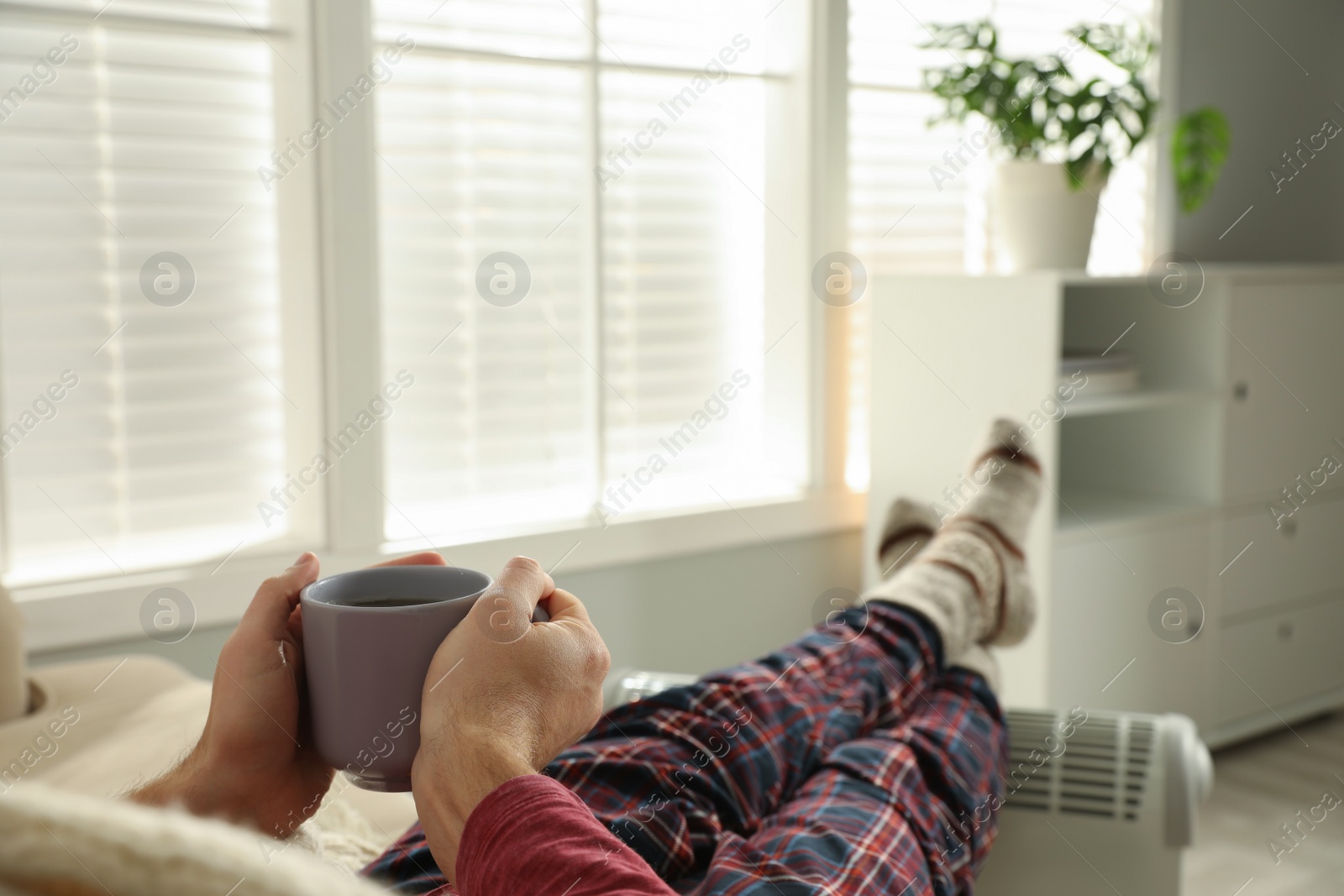 Photo of Man with cup of hot drink warming feet on electric heater at home, closeup. Space for text