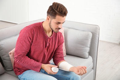 Photo of Young man applying bandage on injured arm at home. First aid