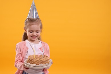 Photo of Birthday celebration. Cute little girl in party hat holding tasty cake with burning candles on orange background, space for text