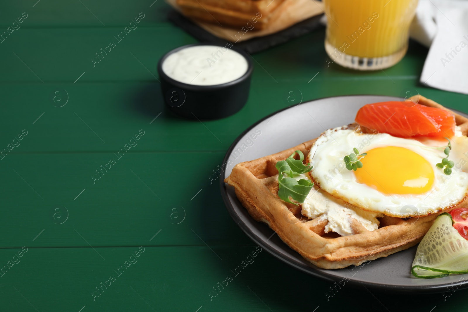 Photo of Delicious Belgian waffle with fried egg, salmon, cream cheese and vegetables served on green wooden table, closeup. Space for text