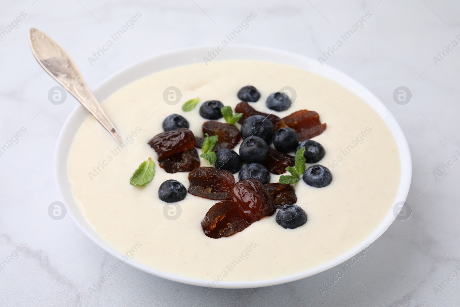 Photo of Delicious semolina pudding with blueberries, dates, mint and spoon in bowl on white table, closeup