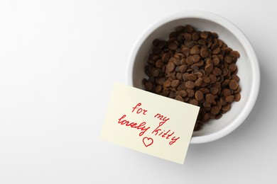 Photo of Feeding bowl with dry cat food and cute note on white table, top view. Space for text