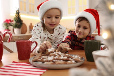 Photo of Cute little children taking tasty Christmas cookies from plate at  table in kitchen