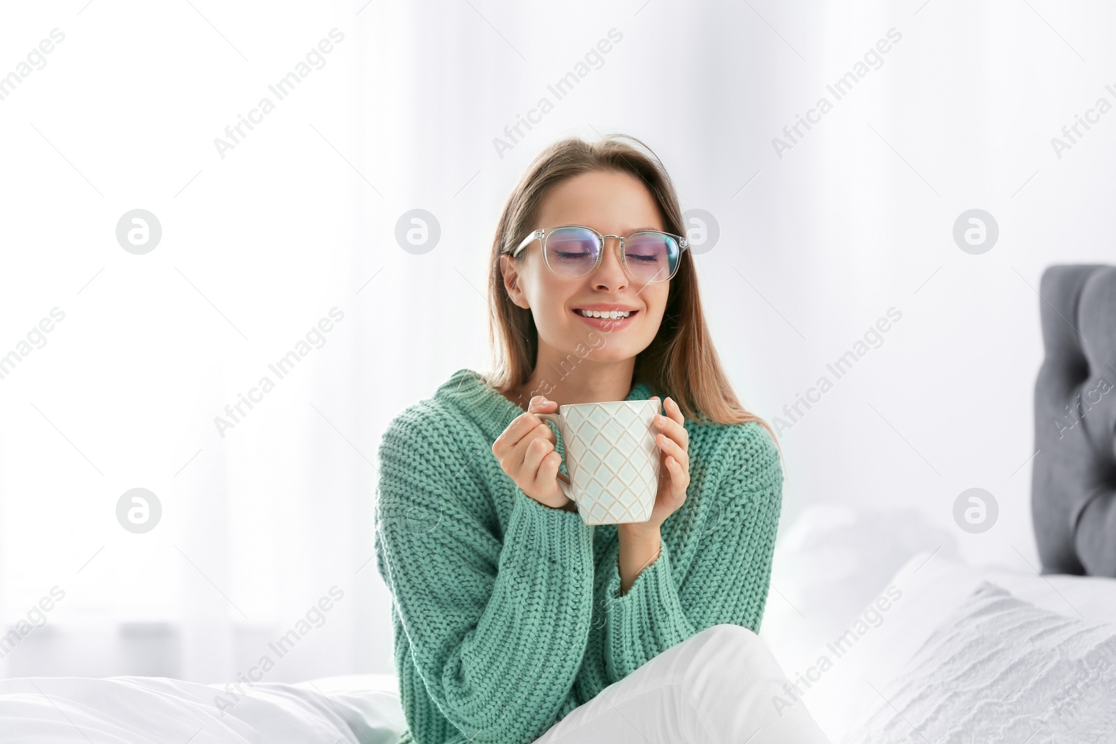 Photo of Young woman in warm sweater with hot drink on bed at home