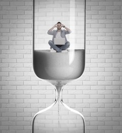 Worried man with laptop sitting inside hourglass against white brick wall. Flowing sand symbolizing coming deadline