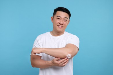 Photo of Handsome man applying body cream onto his elbow on light blue background