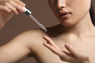 Beautiful young woman applying cosmetic serum onto her finger on brown background, closeup