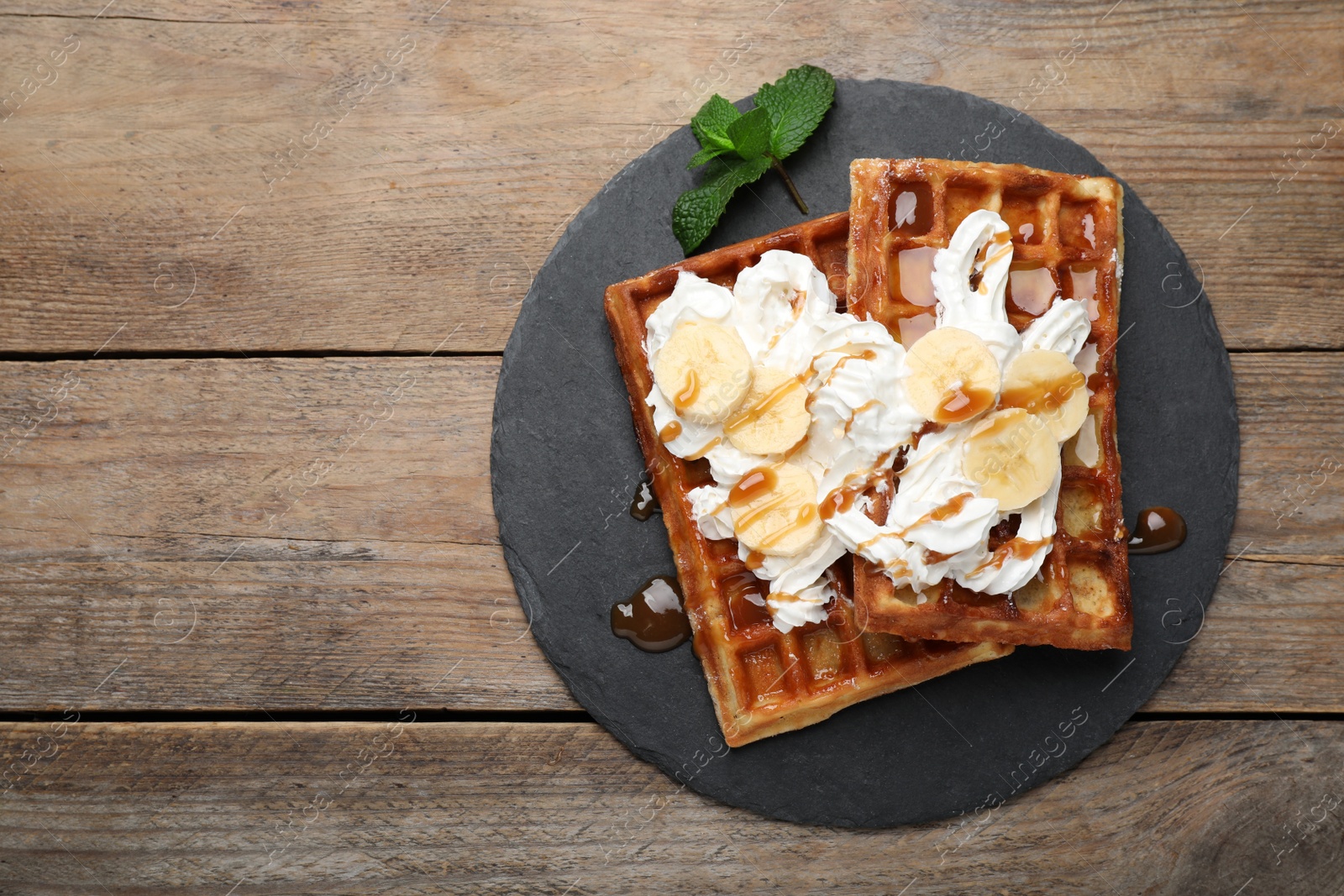 Photo of Slate plate of delicious Belgian waffles with banana, whipped cream and caramel sauce on wooden table, top view. Space for text