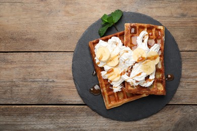 Slate plate of delicious Belgian waffles with banana, whipped cream and caramel sauce on wooden table, top view. Space for text