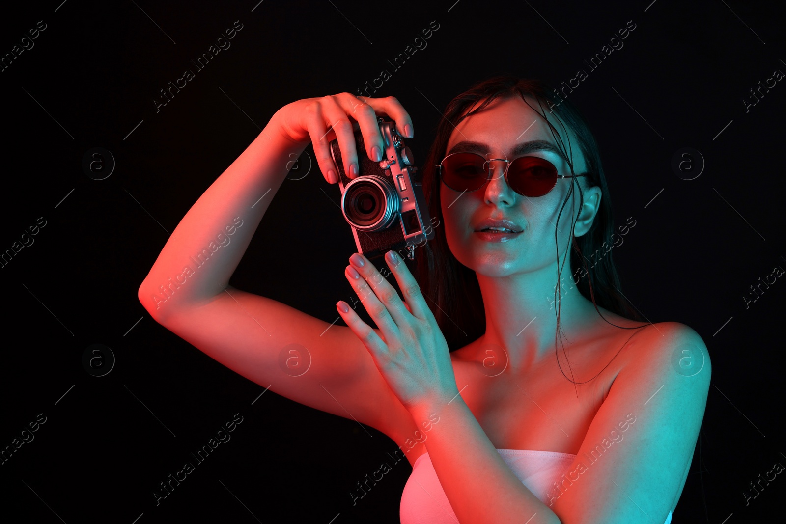 Photo of Beautiful woman with sunglasses and vintage camera posing in neon lights against black background