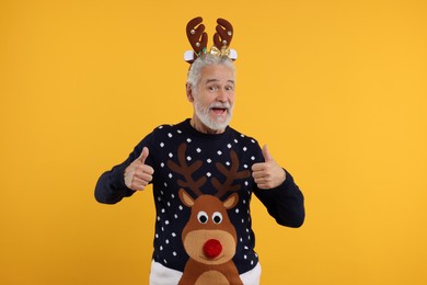 Photo of Emotional senior man in Christmas sweater and reindeer headband showing thumbs up on orange background