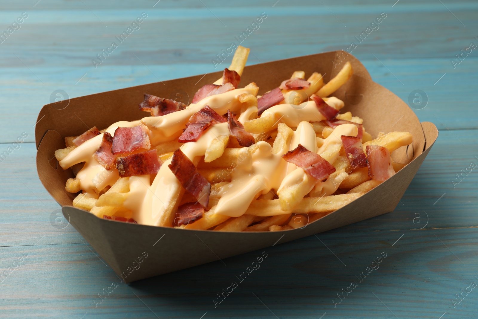 Photo of Tasty potato fries, cheese sauce and bacon in paper container on light blue wooden table, closeup