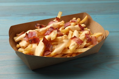 Photo of Tasty potato fries, cheese sauce and bacon in paper container on light blue wooden table, closeup