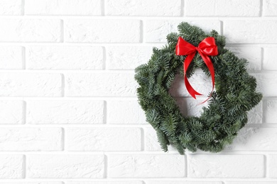 Photo of Christmas wreath made of fir tree branches with red ribbon on white brick wall, space for text