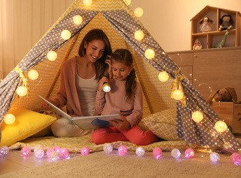 Photo of Mother and daughter with flashlight reading book in play tent