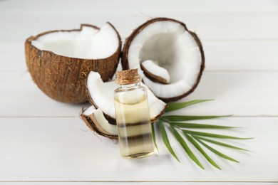 Photo of Bottle of organic coconut cooking oil, leaf and fresh fruits on white wooden table, closeup