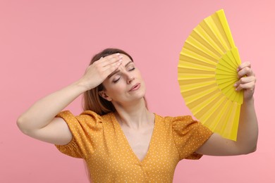 Beautiful woman waving yellow hand fan to cool herself on pink background