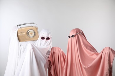 Photo of Glamorous ghosts. Women in color sheets and sunglasses with retro radio receiver on light grey background